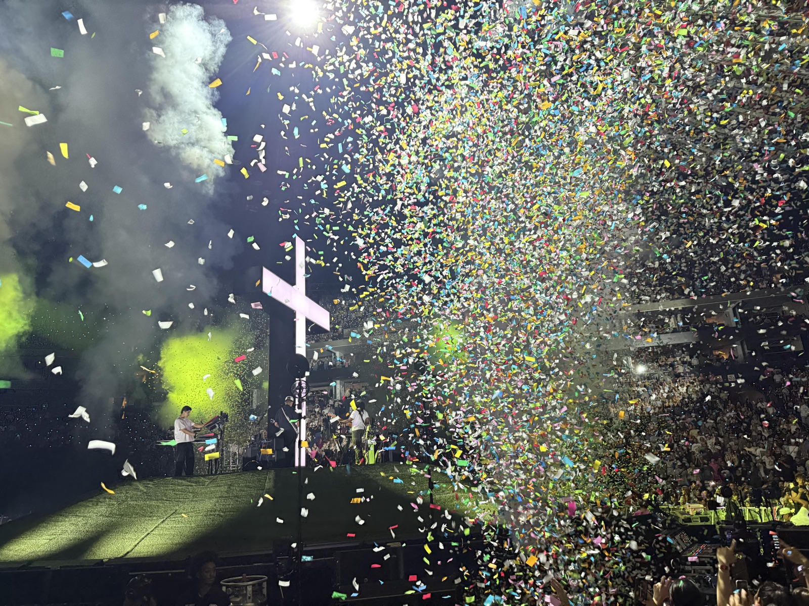 A lit cross on a stadium stage as confetti falls over the crowd